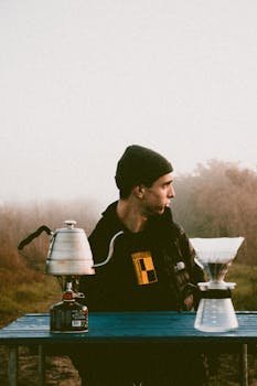 Man makes pour-over coffee outside on a foggy day, enjoying morning freshness.